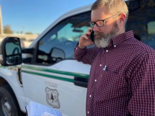 Image of bearded man in plaid shirt talking on cell phone in front of a truck.