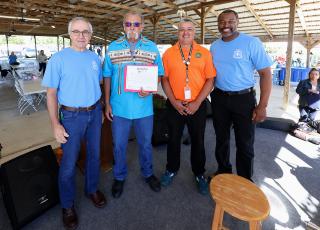 Four people standing under a large outdoor pavilion, posing for a group photo. One person in the center is holding up a certificate.