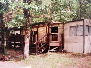 Historic-looking image of tan trailer house with brown wooden deck in the woods.