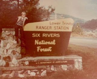 Historic photo of woman sitting on rock wall in jacket, pants and boots next to USDA Forest Service sign that says Lower Trinity Ranger Station, Six Rivers National Forest.