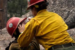 In a rocky forest a firefighter helps to apply eye drops to a fellow firefighter.