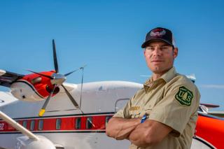 Image shows a man in a Forest Service uniform standing in front of a twin-engine aircraft.