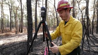 Employee in Nomex and hard hat adjusts laser scanner to measure prescribed burn in experimental forest.