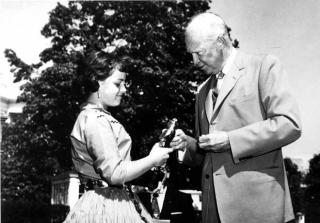 Image shows President Eisenhower handing a young woman a golden statue of Smokey Bear in front of a tree.
