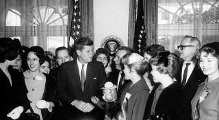 Image shows President Kennedy surrounded by young women, one of whom is holding a doll of Smokey Bear.