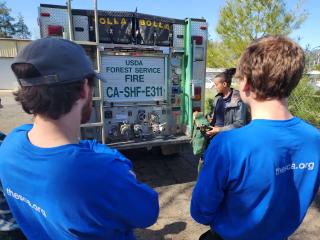 Two interns with their backs facing the camera listen to an instructor at the back of a fire engine as part of their wildland fire training academy course.