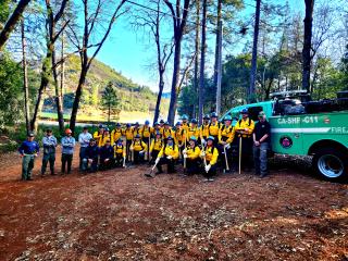 Students in fire protective clothing holding tools like Pulaskis gather with instructors near Forest Service truck in front of Shasta Lake.