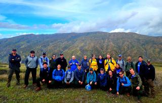 Group photo: Staff ride participants, including Mendocino Hotshots and student interns, pose for a photo. In background, a hill; above, blue skies with white, fluffy clouds.