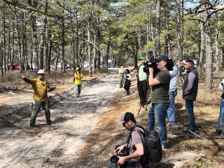 A group of people (on the right) photographinc two men dressed in firefighter uniform (on the right) out in the forest