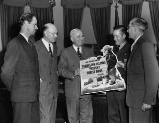 Image shows President Harry Truman standing in the Oval Office holding a poster of Smokey Bear with four other men.