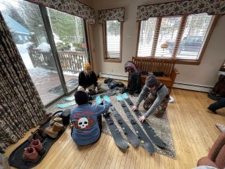 Participants sit on a wood floor with skis spread out among them.