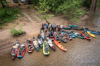 Drone photo from above: Kayaks, canoes and inflatable rafts all full of trash, pulled up to river banks. Standing on shore are volunteers who conducted the river cleanup.