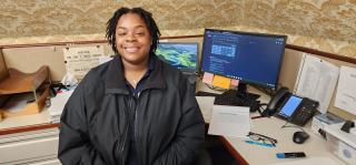 A'Precious Norfleet, wearing a black jacket, sits facing away from her desk and smiling at the camera.