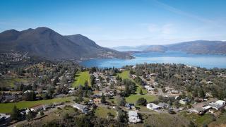 Landscape image of houses surrounded by green trees with large lake and mountains in the background.