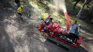 Aerial view looking down on a red piece of equipment being fed tree branches by firefighters in yellow.