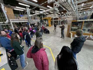 Large group stands in large room with steel beams and concrete floor. They are listening to a lab employee who is wearing a headset microphone. 