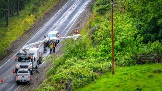 White utility trucks along rural road inspect a powerline next to a forest.