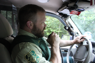 Image shows a man in a law enforcement uniform driving his car, answering a radio inquiry by holding the receiver up to his mouth.