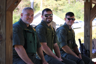 Three men in law enforcement uniforms leaning against a fence smile at the camera.