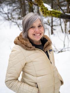 Barb Miranda in a white down coat outdoors in the snow. A moss-covered branch is just behind her head and forest is in the background.