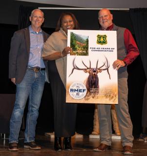 On stage, Associate Chief Angela Coleman flanked by two men from Rocky Mountain Elk Foundation. Associate Chief and man to her left are jointly holding a large foam-board poster. Image on poster: large elk in prairie; inset upper left, photo of Kootenai National Forest. Upper right: Forest Service insignia; lower left, foundation logo. Text: For 40 years of conservation.