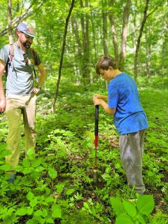 Standing in forest, a professor watches a middle school student use a tool to take a soil sample.