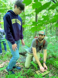 In forest, assistant professor kneels down over a tool to collect a soil sample while a student in a West Virginia sweatshirt stands next to him, observing.