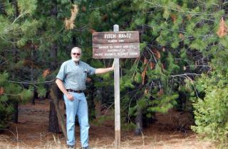Roger Stutts in forest. He's casually posed with his left hand on a wooden sign that says Fitch Rantz, planted 1984, Institute of Forest Genetics, Placerville, California, Pacific Southwest Research Station.