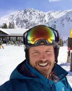 A man in skiing gear smiling at the camera in front of a mountain ski resort.