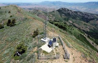 A view from a drone of communication equipment on concrete pads, enclosed by a chain link fence, on top of a mountain ridge.