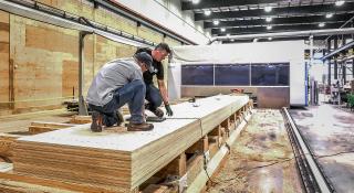 Two people kneeling on top of a stack of engineered wood panels inside a factory.