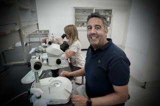 A man sits smiling in front of a microscope. A woman peers into a microscope at a stool behind him. They are in a laboratory.