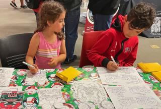 Two children with markers in their hand sit at a table with coloring books.