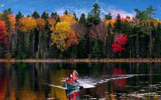 Two people in a canoe crossing a calm lake, with a lush fall forest in the background.