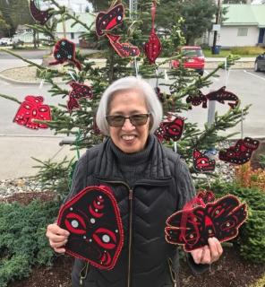 Maryanne Rainey standing outside, in front of a decorated pine tree, holding home made ornaments in both hands.