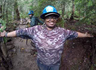 A person wearing a hard hat, standing on a forest trail with arms outstretched to her sides as if embracing the photographer.