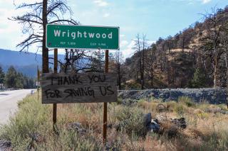 A highway sign when entering the town of Wrightwood, with a plywood sheet nailed to the bottom with "THANK YOU FOR SAVING US" written with black spray paint.