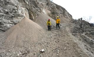 Two people in yellow jackets wearing red and yellow hard hats stand holding shovels next to large piles of gravel (gravity-induced erosion).