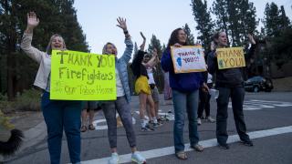 A line of people hold signs and wave by the road expressing their thanks for firefighters.