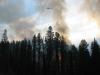 A picture of a wildfire burning in a heavily forested area with a helicopter and water bucket attached to the helicopter seen flying above the fire. 