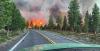 A raging forest fire viewed through the front windshield of a vehicle on a paved road.