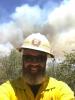 A bearded man in a wildland firefighter uniform takes a selfie in front a wooded area covered in smoke.