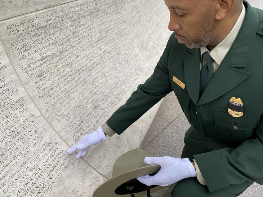 A man in Forest Service LEI uniform, kneeling down in front of a marble wall full of names of fallen law enforcement officers and, holding his hat on his left hand and with his right while wearing white gloves, points at a particular name on the wall list.