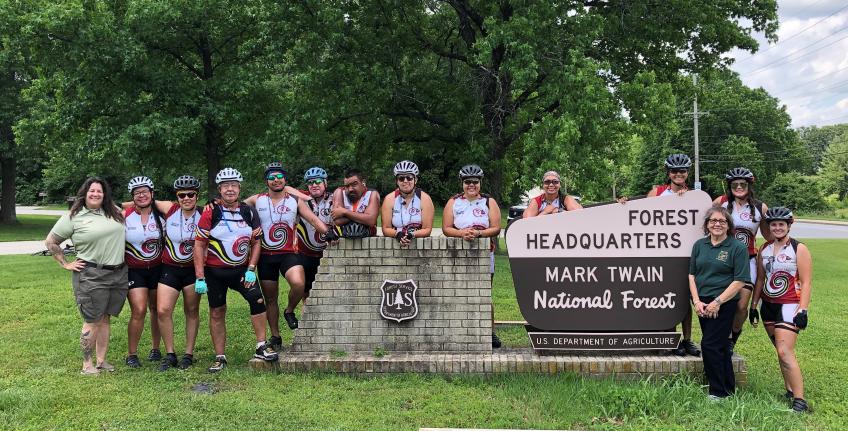 Group photo: Bicyclists wearing helmets with two Forest Service employees grouped around forest headquarters sign for Mark Twain National Forest.