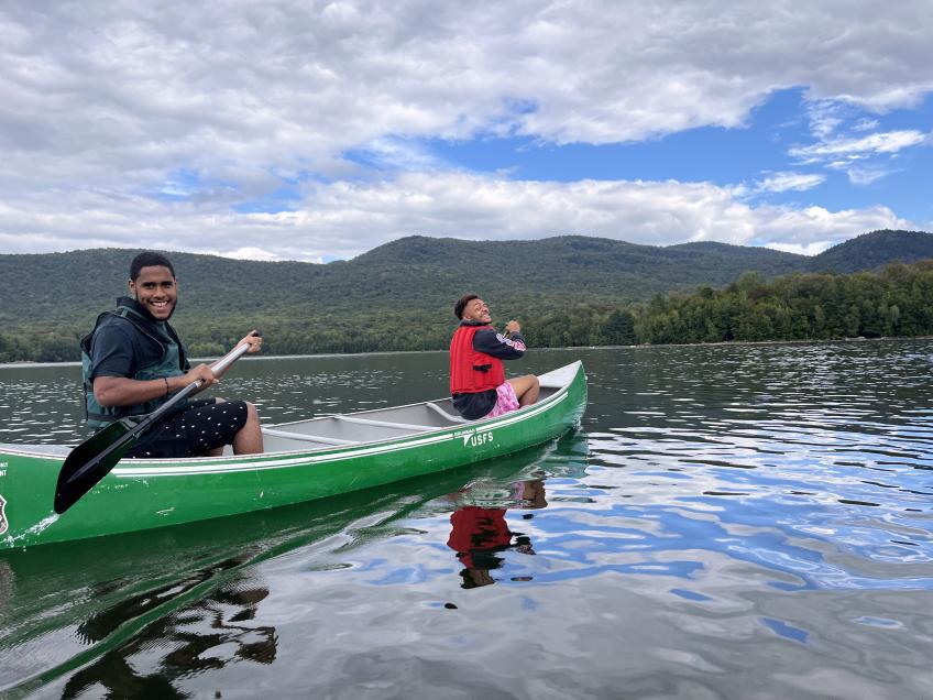 Two students twist back to look at the camera from their canoe on a reservoir with forest in background. The blue sky and fluffy clouds reflect in the water.