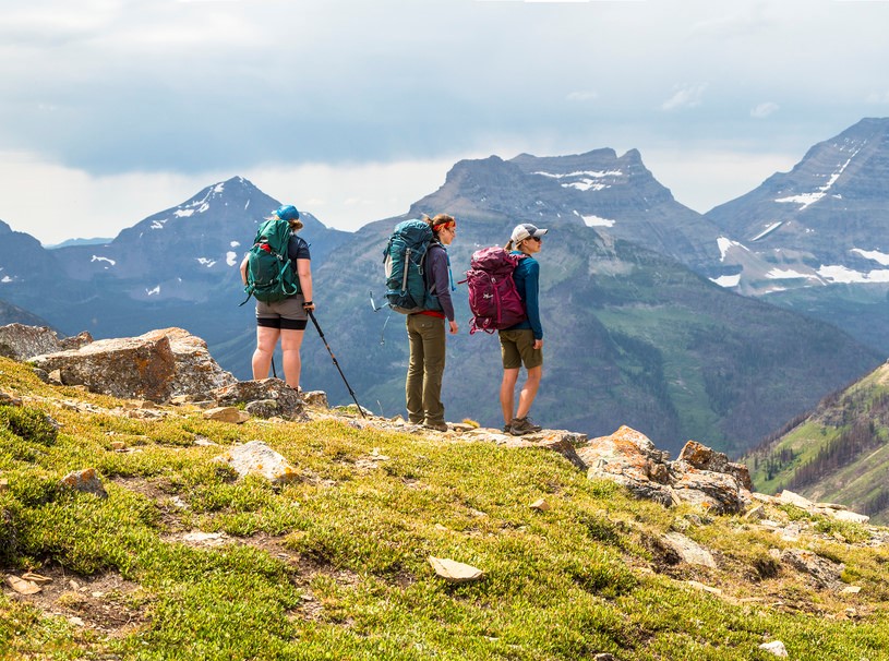 "A picture of three hikers with large backpacks standing on a mountain ridge. They are looking away from the camera at a backdrop of jagged, snowy mountain peaks."