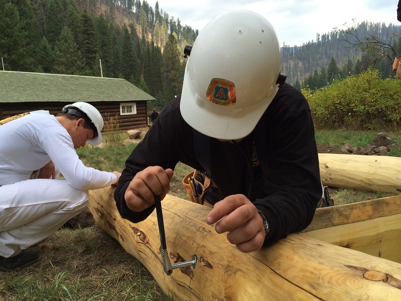 Two youth in hard hats repair a cabin in the forest.