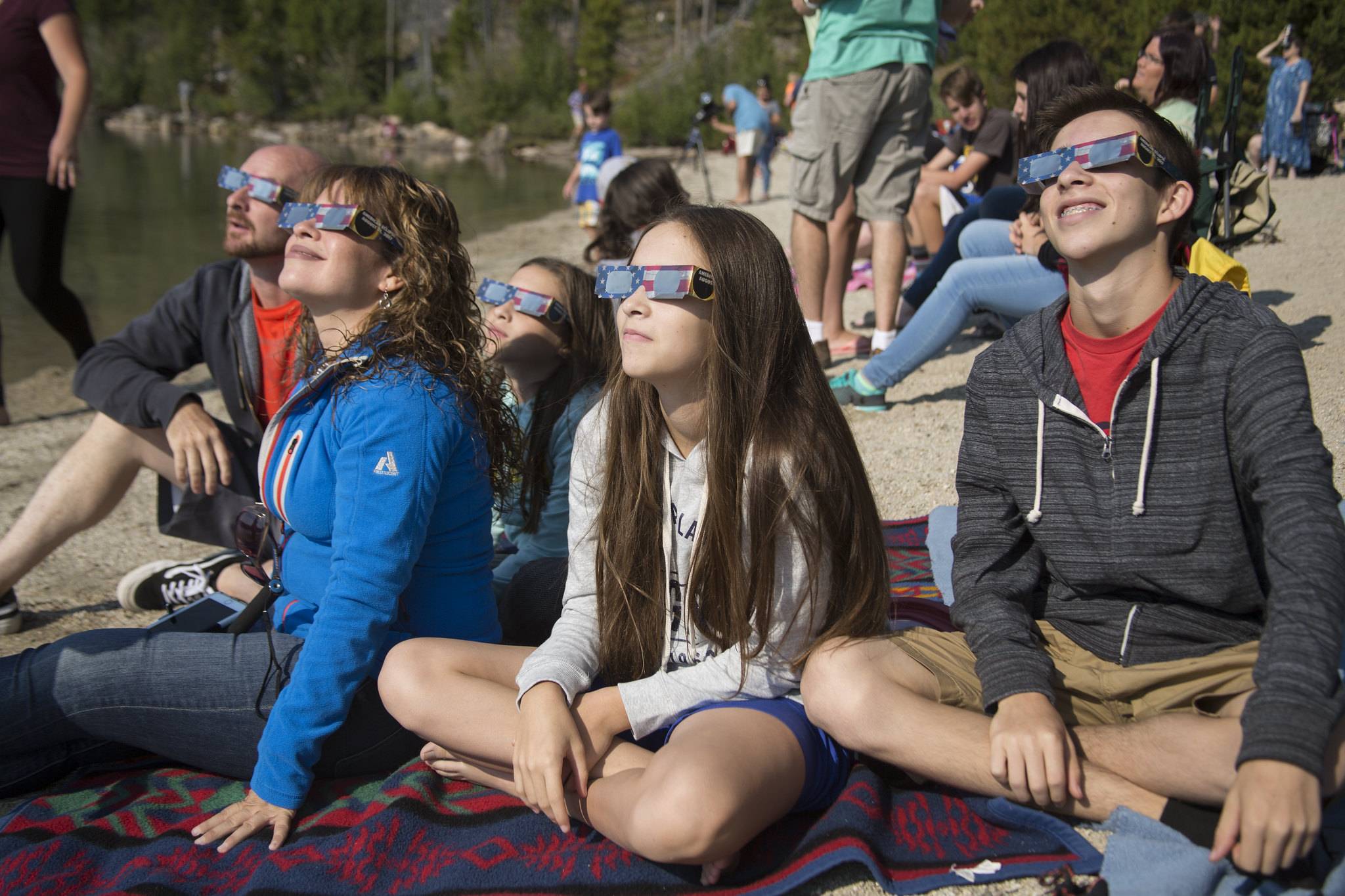 A family of five sits on the ground wearing solar eclipse glasses while looking up. A crowd sits in the background.