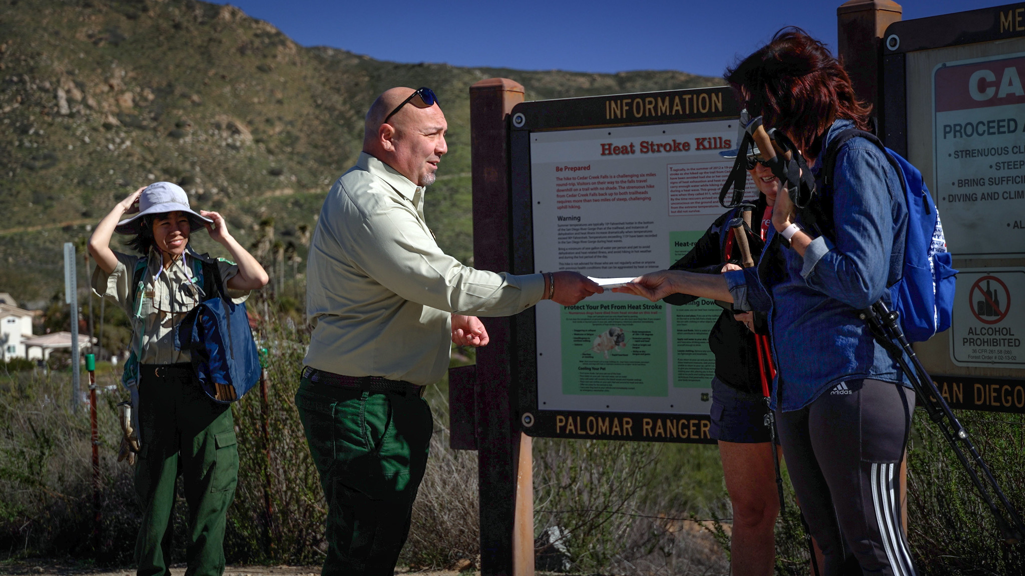 A public affairs officer speaks to 2 hikers at a trailhead. Another employee observes.