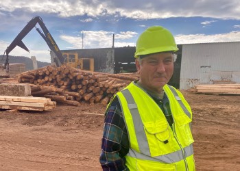A man in a hard hat and vest stands in front of heavy equipment and a timber pile